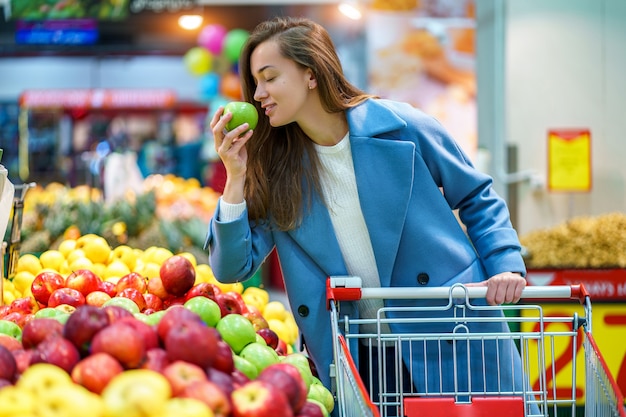 Portrait of a smiling happy attractive young woman buyer with cart in the grocery store during choosing and buying fresh apples at fruit department