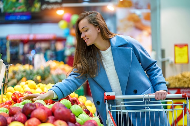 Ritratto di un compratore attraente felice sorridente della giovane donna con il carrello nel supermercato della drogheria durante la scelta e l'acquisto delle mele fresche al dipartimento della frutta
