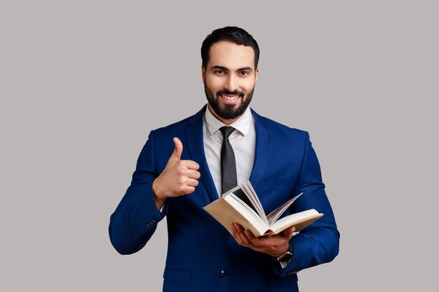 Portrait of smiling happy bearded man showing thumbs up gesture holding and reading book, likes genre and plot, wearing official style suit. Indoor studio shot isolated on gray background.