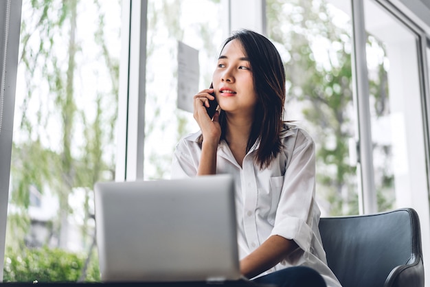 Portrait of smiling happy beautiful asian woman relaxing using technology of smartphone and looking in window while sitting on chair.