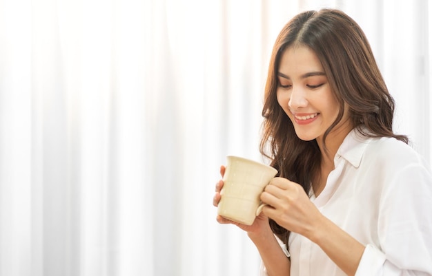 笑顔の幸せな陽気な美しさの肖像画かなりアジアの女性リラックスして飲んでホットコーヒーやお茶を見て女の子の伐採は、自宅のベッドで休日の朝の休暇で朝食を楽しんでいます