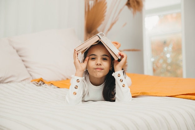 Портрет улыбающейся счастливой маленькой девочки, держащей книгу на голове и лежащей на кровати.