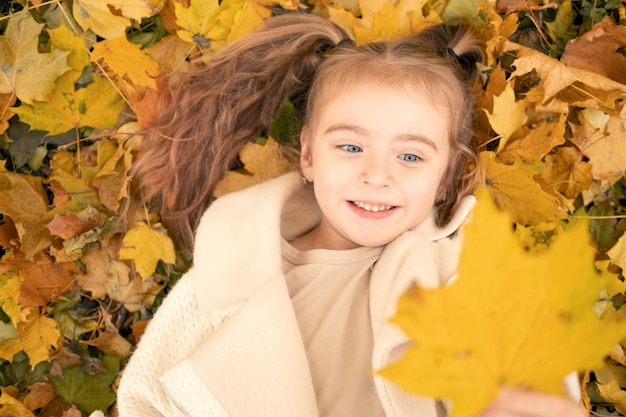 portrait smiling happy little girl lying on grass in nature in autumn with yellow maple leaves