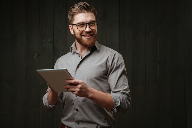 Портрет улыбающегося счастливого человека в очках, держащего планшетный компьютер и смотрящего в сторону, изолированного на черной деревянной поверхности