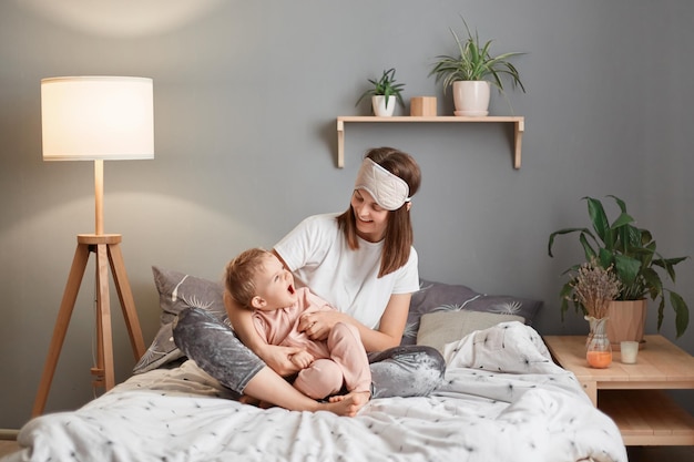 그녀의 이마에 수면 마스크에 웃는 행복한 어머니의 초상화와 행복을 표현하는 그녀의 흥분된 유아 딸을 안고 집에서 침대에서 노는 아기의 초상화