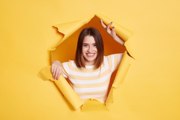 Portrait of smiling happy woman stands in torn paper hole expressing positive emotions being in good mood looking through breakthrough of yellow background