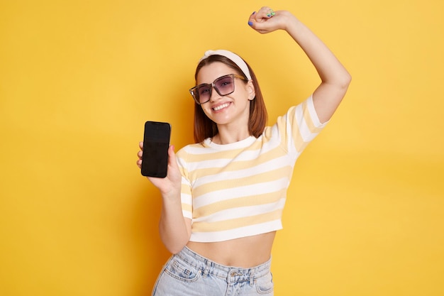 Portrait of smiling happy woman wearing striped shirt posing isolated over yellow background female celebrating her success raising hand showing cell phone with space for advertisement