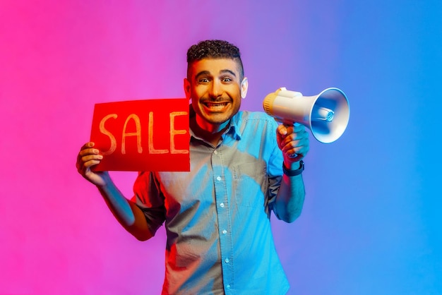 Photo portrait of smiling happy young adult handsome man in shirt holding megaphone and big sale, black friday shopping. indoor studio shot isolated on colorful neon light background.