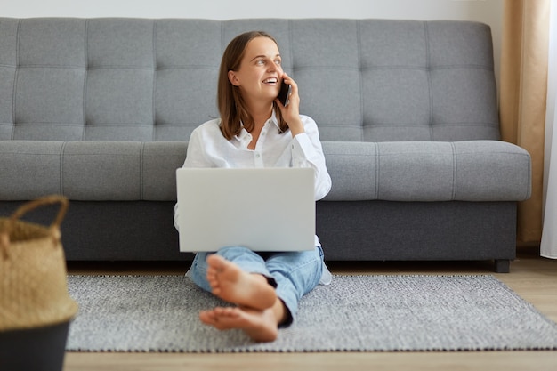 Portrait of smiling happy young woman using laptop and cellphone while sitting near sofa at home, looking away, having break on work and talking with friend via smartphone.