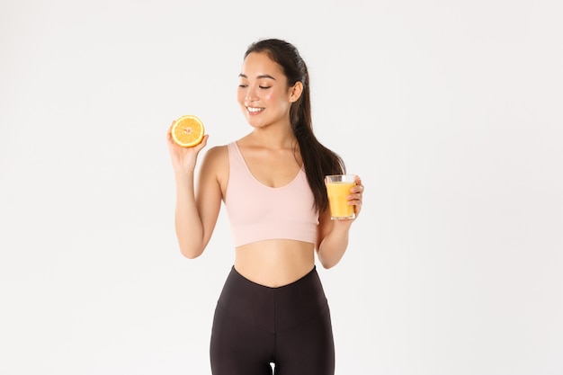 Портрет улыбающейся здоровой и стройной азиатской девушки советует есть здоровую пищу на завтрак, набирать энергию для тренировки, держать свежий сок и апельсин.