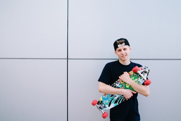 Portrait of a smiling hipster with a longboard in his hands against a gray wall. young man with a skateboard poses on a gray