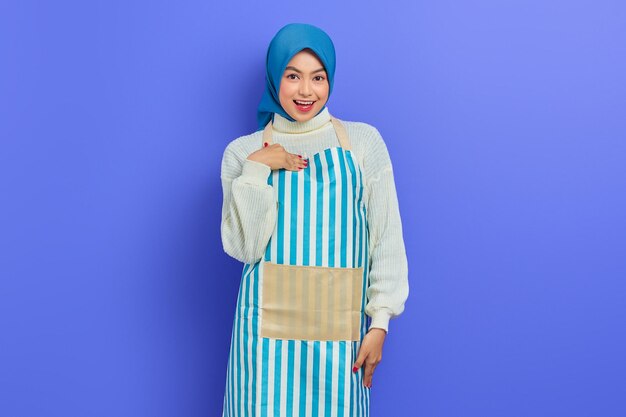 Портрет улыбающейся домохозяйки в хиджабе и фартуке, указывающей на себя, гордо изолированной на фиолетовом фоне Концепция мусульманского образа жизни домохозяйки