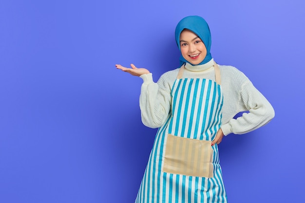 Portrait of smiling housewife woman in hijab and apron showing product with hand isolated on purple background People housewife muslim lifestyle concept