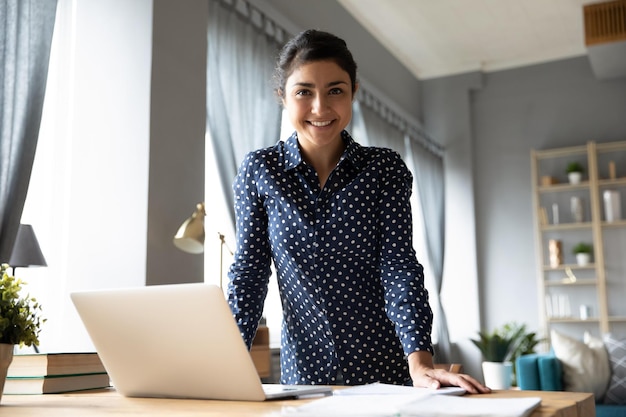 Portrait smiling Indian girl standing at desk with laptop looking at camera successful businesswoman freelancer posing for photo at workplace satisfied excited female student in living room