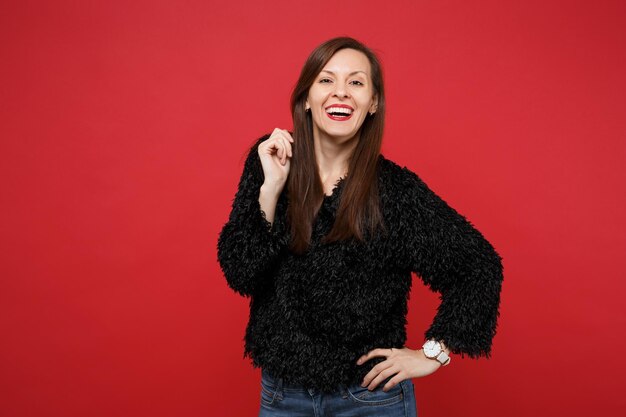 Portrait of smiling joyful beautiful young woman in black fur sweater standing isolated on bright red wall background in studio. People sincere emotions, lifestyle concept. Mock up copy space.