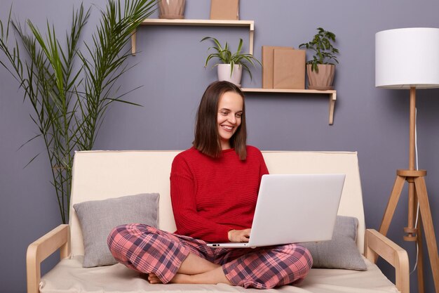 Portrait of smiling joyful woman freelancer with dark hair wearing home clothing sitting on sofa in living room female working on laptop typing on keyboard journalist writing article