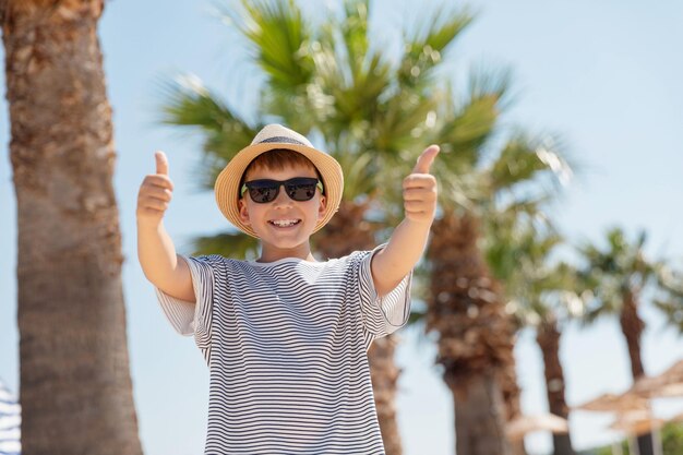 Portrait of smiling kid boy with in straw hat and sunglasses showing thumb up staying outdoor with palm trees and sky on the background looking at the camera