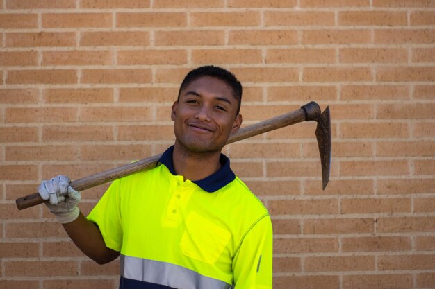 Portrait of a smiling laborer with a hoe over his shoulder looking at camera, brick wall on the background