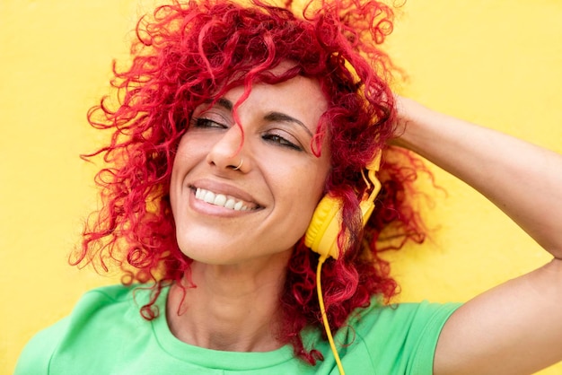 Portrait of a smiling latin woman with red afro hair wearing a
green tshirt looking at the horizon and listening to music with
yellow headphones over a yellow background