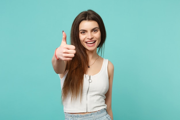 Portrait of smiling laughing young woman in light casual clothes showing thumb up isolated on blue turquoise wall background in studio. People sincere emotions lifestyle concept. Mock up copy space.