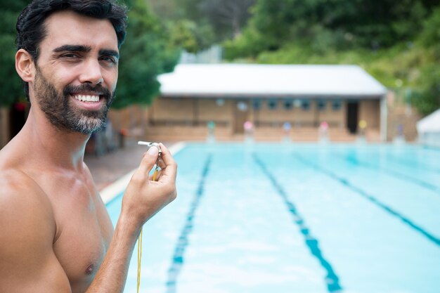 Portrait of smiling lifeguard standing with whistle near poolside