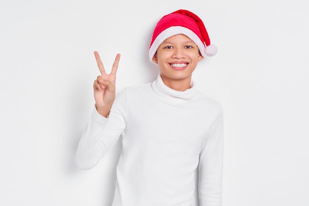 Portrait of smiling little Asian man in christmas hat showing peace or victory sign isolated over white background Happy New Year 2023 holiday concept