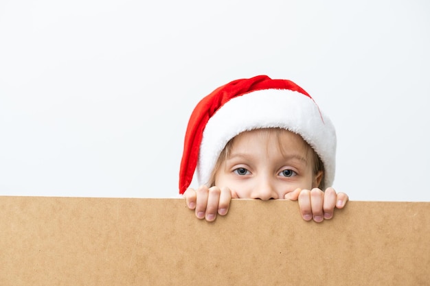 Portrait of smiling little girl with Christmas hat. Kid peeping behind blank holidays board. Happy cute child posing behind kraft paper panel, isolated on white background.
