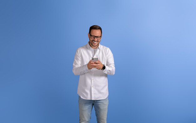 Foto ritratto di un imprenditore sorridente che manda un messaggio online tramite telefono cellulare su uno sfondo blu isolato