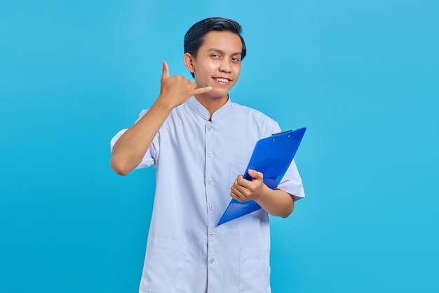 Portrait of smiling male nurse holding clipboard standing and making call gesture on blue background