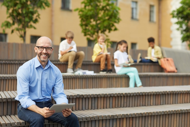 Portrait of smiling male teacher looking at camera while holding digital tablet sitting on bench outdoors with group of schoolchildren in background, copy space