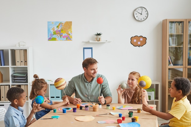 Ritratto dell'insegnante maschio sorridente che lavora con un gruppo multietnico di bambini che dipingono i modelli del pianeta nella lezione di arte e artigianato