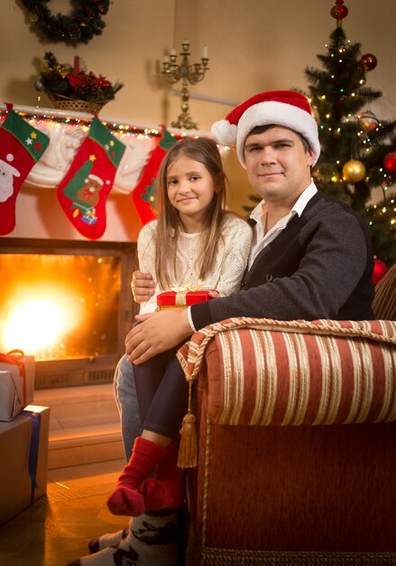 Portrait of smiling man and girl relaxing on sofa at fireplace at Christmas eve