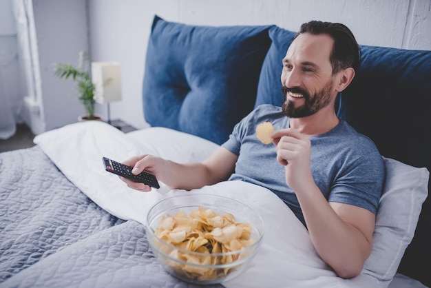Photo portrait of smiling man watching tv and eating chips while resting in the bed at home