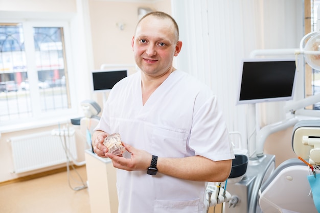 Portrait of smiling man in white doctor robe uniform is standing in office, clinic with chair, instruments. Dentist workplace. Orthodontist is preparing for visit of patient. Dentistry concept.