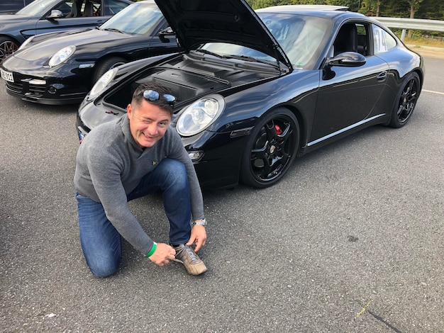 Portrait of smiling mature man winking while tying shoelace on road against car