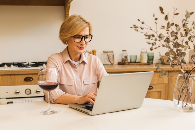 식탁에서 노트북을 사용하는 동안 와인 한 잔을 들고 웃고 있는 성숙한 고위 여성의 초상화. 집 개념에서 일하는 프리랜서.