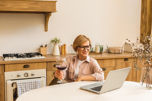 台所のテーブルでラップトップを使用しながらワインのグラスを保持している笑顔の成熟した年配の女性の肖像画。フリーランス在宅勤務のコンセプト。
