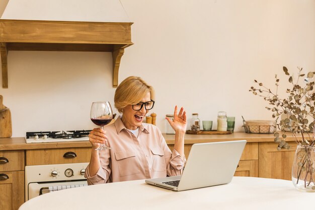 Portrait of smiling mature senior woman holding glass of wine while using laptop on kitchen table. Freelance working at home concept.