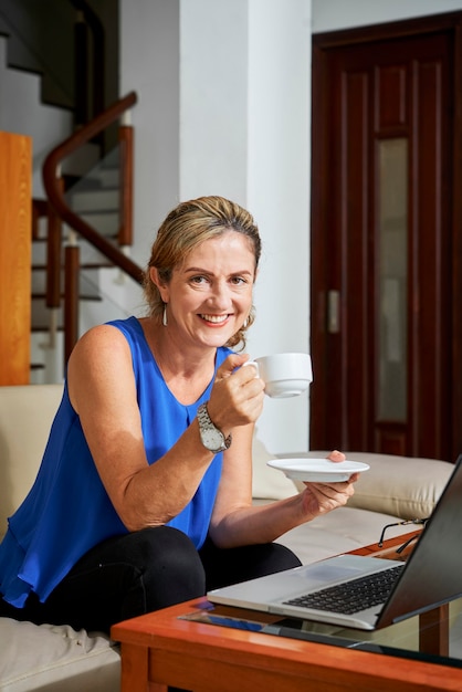 Portrait of smiling mature successful woman drinking tea and working on laptop at home due to coronavirus pandemic