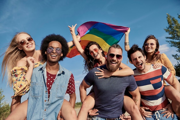 Photo portrait of smiling men with rainbow flag carrying friends against sky