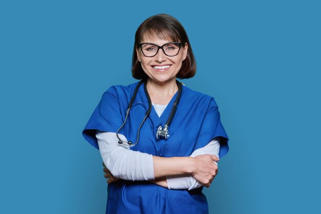 Photo portrait of smiling middle aged doctor with stethoscope looking at camera on blue studio background confident woman in glasses with arms crossed medicine health care staff medical service concept