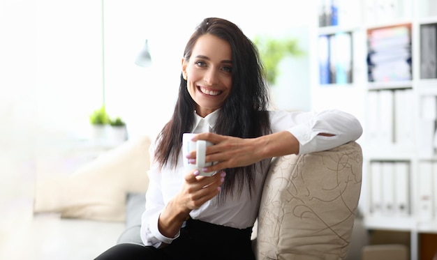 Portrait of smiling minded successful employee business woman in white shirt with cup Concept of career achievement and rest in workplace