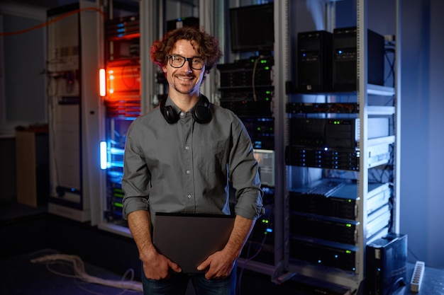Портрет улыбающегося сетевого инженера, стоящего с ноутбуком в серверной комнате