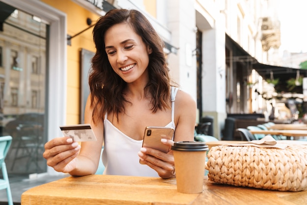 Portrait of smiling nice woman with straw bag holding credit card and using cellphone while sitting in cozy cafe outdoors