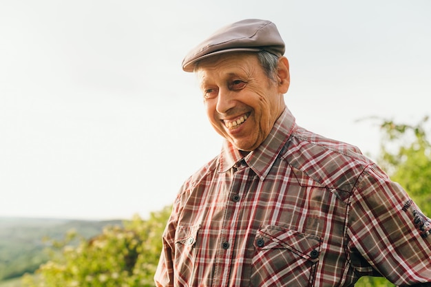 모자와 셔츠를 입고 옆을 바라보고 웃고 있는 웃는 노인의 초상화 야외에서 행복한 은퇴한 할아버지 초상화 은퇴한 농부의 초상화