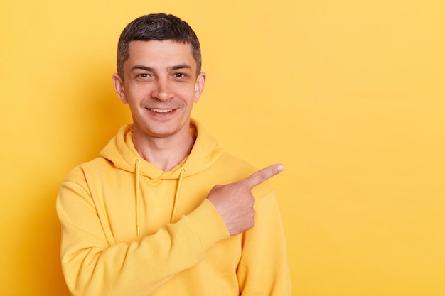 Portrait of smiling optimistic young adult man wearing casual style hoodie posing isolated over yellow background standing and pointing finger aside showing advertisement area