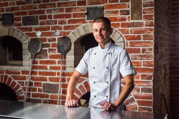 Photo portrait of a smiling pizza chef standing on the background of a woodburning oven in the kitchen