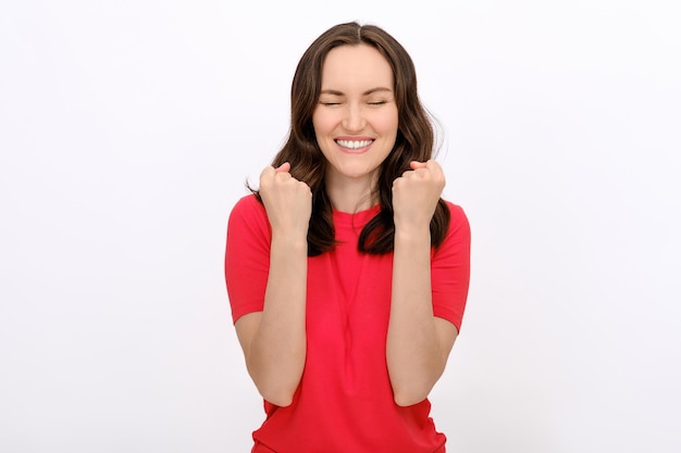 Ritratto di donna bruna soddisfatta sorridente con gli occhi chiusi in maglietta rossa con braccia piegate e pugni in alto su sfondo bianco per la pubblicità mostra modello di spazio copia concetto di successo fortuna