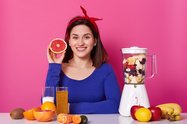 Portrait of smiling positive female holding cut fruit in her hand. Young model waits for smoothie to be ready. Cheerful happy woman stick to healthy diet with pleasure. Healthy lifestyle concept.