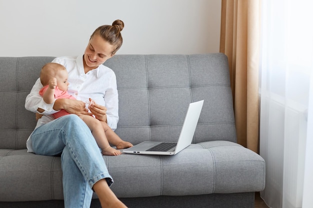 Portrait of smiling positive young mother with baby woman with bun hairstyle wearing white shirt and jeans playing with toddler kid and working on computer